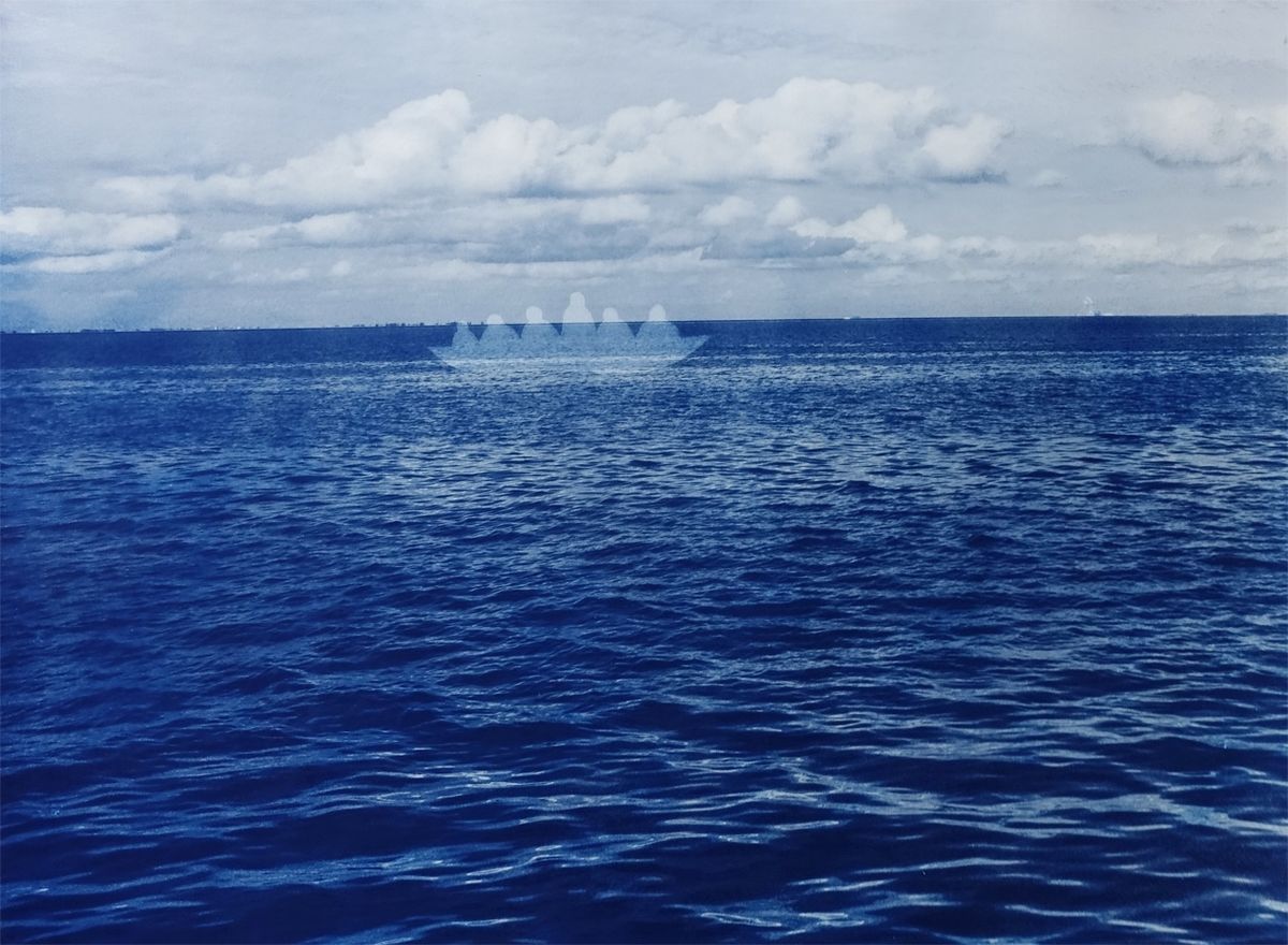 Cyanotype print of a photographic seascape with rippling deep blue water and a cloudy sky, with a faint pale silhouette of a small boat carrying a row of human figures on the horizon.