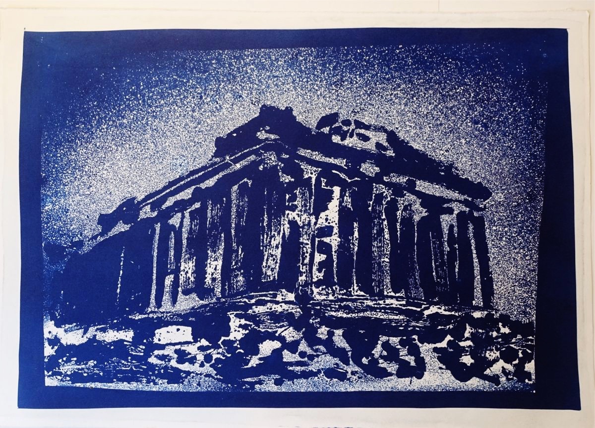 Cyanotype print in deep blue showing a ruined Doric temple seen from a low corner angle, columns and entablature in loose brushy strokes over a rocky base, against a fine white speckled sky, framed by a solid blue border on white paper.