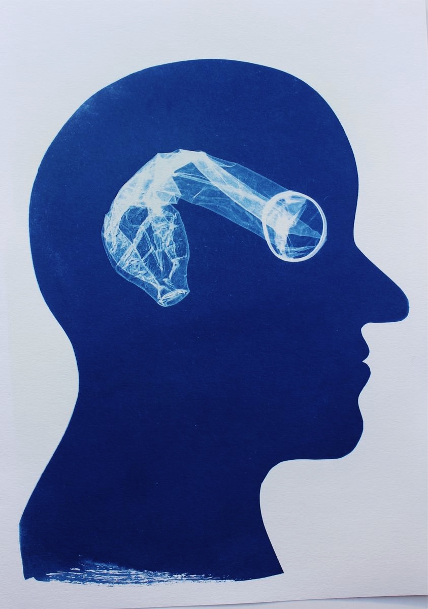 Cyanotype print of a bald human head in blue profile silhouette facing right on white paper, with the translucent white imprint of a crumpled condom placed horizontally inside the head at eye level.