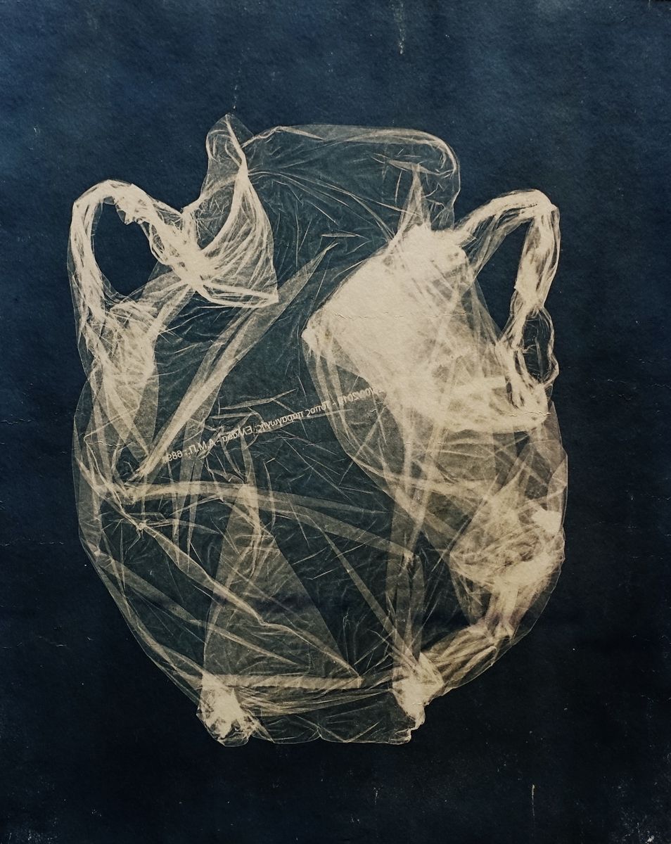 Toned cyanotype print in dark blue-black and warm cream showing the translucent imprint of a crumpled plastic bag whose two handles and rounded body form the shape of a two-handled vessel, with faint printed text visible across the plastic.