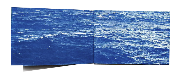 Open blue cyanotype booklet spread showing a continuous image of a rippling ocean surface stretching across both pages.