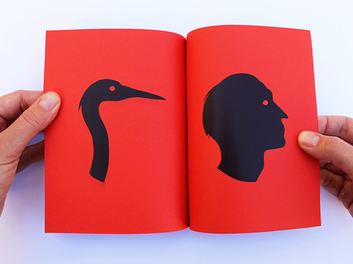 Two hands holding open a red booklet spread showing a black silhouette of a bird's long curved neck and head on the left page, facing a black silhouette of a human head in profile on the right page.