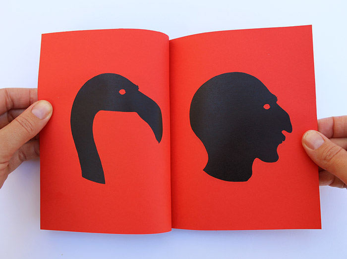 Two hands holding open a red booklet spread showing a black silhouette of a flamingo's curved neck and beak on the left page, facing a black silhouette of a human head with an open mouth in profile on the right page.