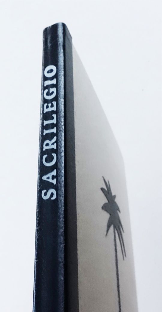 Close-up of a hardbound book standing upright, with a dark textured spine printed with the title in white capital letters and a grey cover showing a black palm tree silhouette.