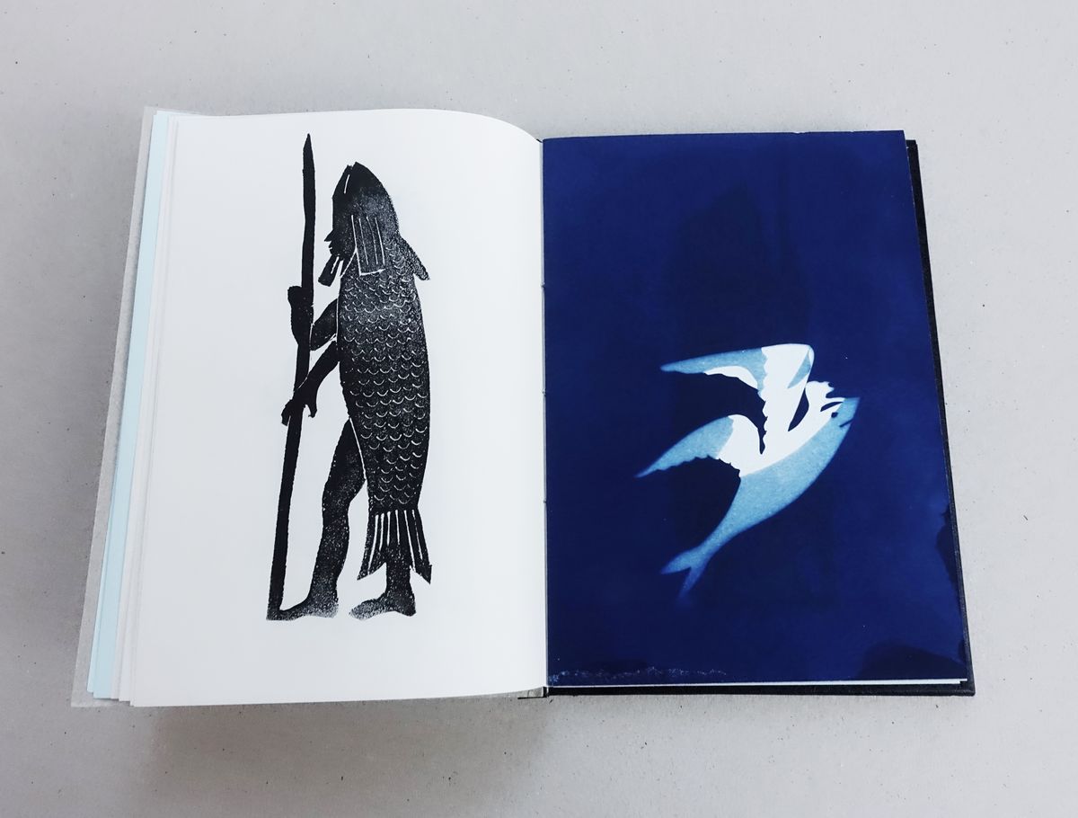 Open book on a grey surface; the left page shows a black block print of a standing figure wearing a large fish costume and holding a tall spear, and the right page a deep blue cyanotype with a pale bird in flight.