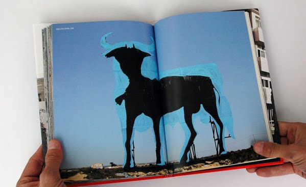Open art book spread with two hands holding it open, showing a black silhouette of a horned goat mural photographed against a blue sky and low buildings.
