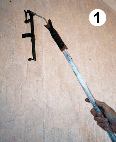 Close-up of a hand holding a long pole tool with a hooked end near a wall-mounted bracket, numbered "1" in the corner as an instructional step.