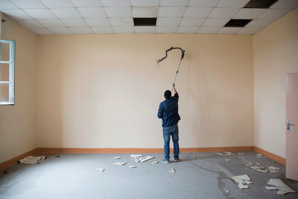 A person standing in an empty room using a long pole tool to draw or mark the wall, with torn debris scattered across the floor.