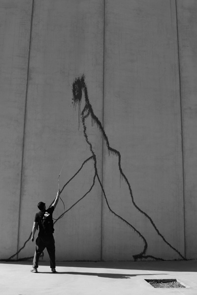 A person using a long tool to paint or mark a tall concrete wall, creating a dark dripping silhouette resembling a walking figure with elongated limbs.