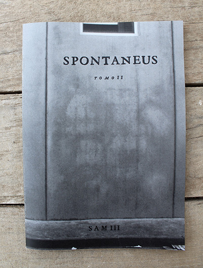 Grey booklet cover titled "SPONTANEUS TOMO II, SAM III" photographed on a wooden surface, showing a faint photographic image of a wall with a dark band across the bottom.
