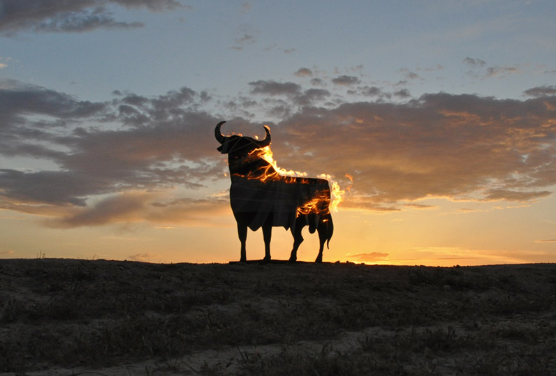 Black silhouette of a large bull-shaped billboard on a hilltop at sunset, flames digitally overlaid across its body, glowing orange and pink clouds filling the sky behind it, in Fortuna, Murcia, Spain.