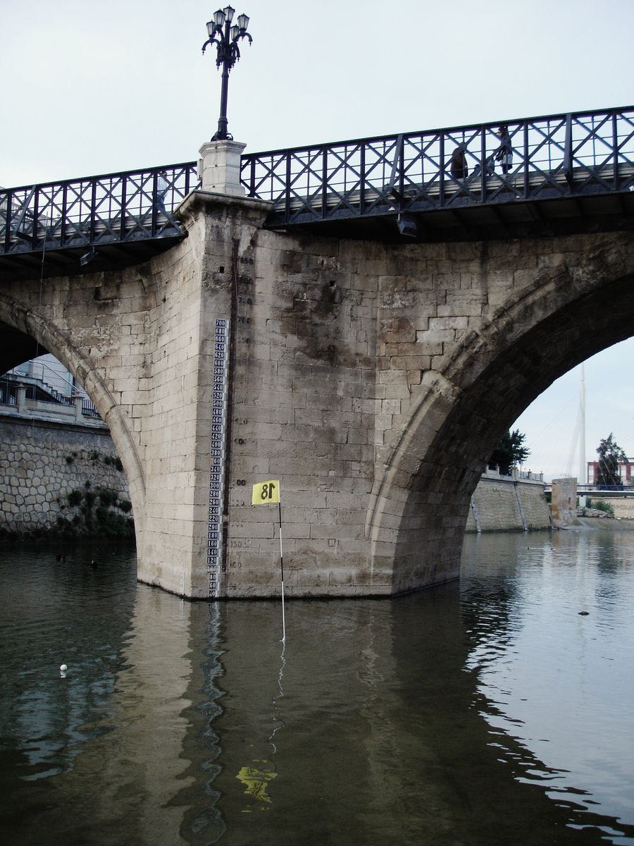 Stone pier of a river bridge with a water-level measuring scale carved into the stone, a small yellow flag marked '18' planted at the waterline, a lamppost and pedestrians crossing the bridge above.