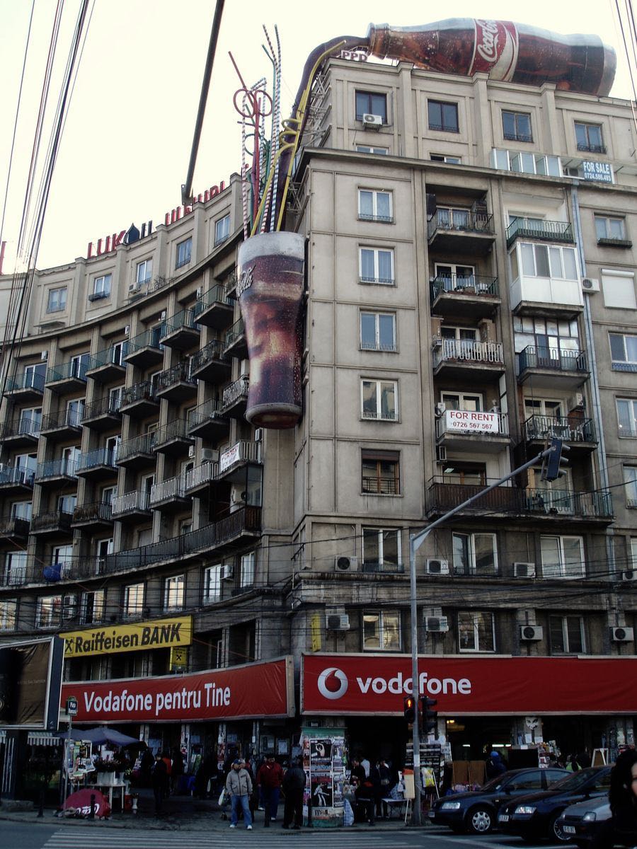 Curved building facade with an oversized Coca-Cola bottle and glass mounted on the rooftop and side wall, bank and phone company signage below.