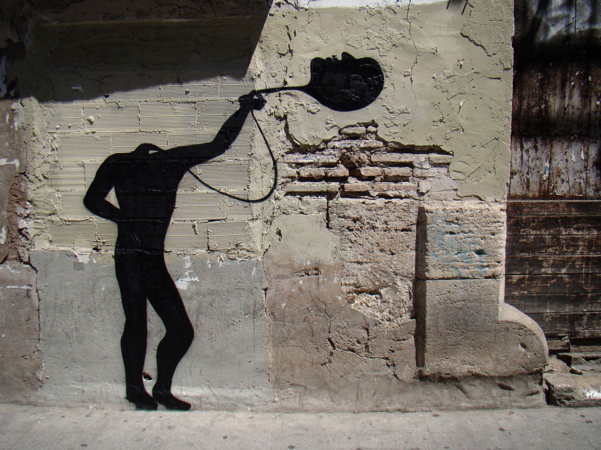 Black silhouette of a bent-over figure with one arm stretched overhead holding a flat oval shape resembling a face or plate, painted on a weathered brick and plaster wall.
