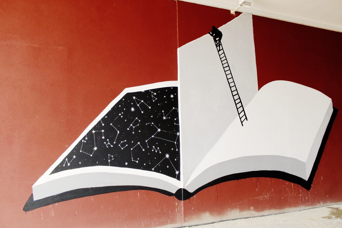 Red wall mural of a giant open book, one page showing a black night sky full of constellations, the other page blank with a black ladder and a small silhouette figure climbing up it.