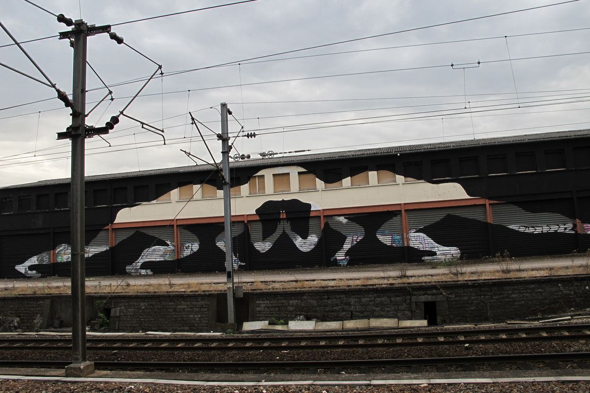Long dark train shed roofline painted with a black silhouette train carriage shape blending into the real roof, railway tracks and overhead power lines in the foreground.