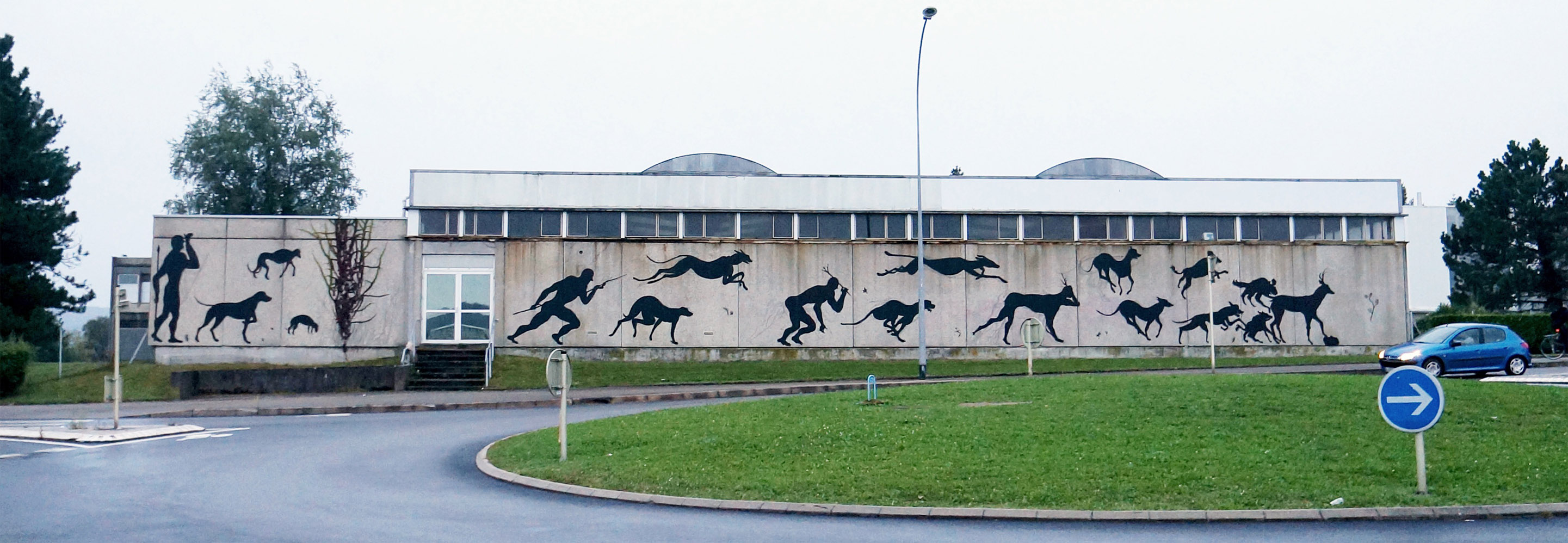 Long low building facade painted with a running black silhouette hunter figure chased by leaping hounds and deer across several panels, two children standing on the grass in front.