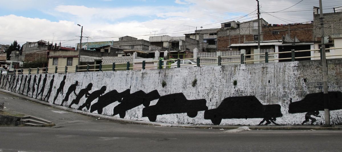 Long mural on a curved retaining wall showing a repeated black silhouette of a car being pushed by a running figure, a caravan-like procession stretching along the wall above a street, with residential buildings visible behind the railing.