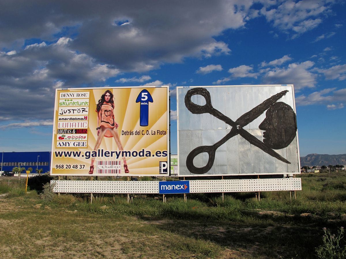 Roadside billboard painted with a black silhouette of a police baton or club shape, an abandoned farmhouse and palm tree beside it, a corrugated metal fence running along the background.