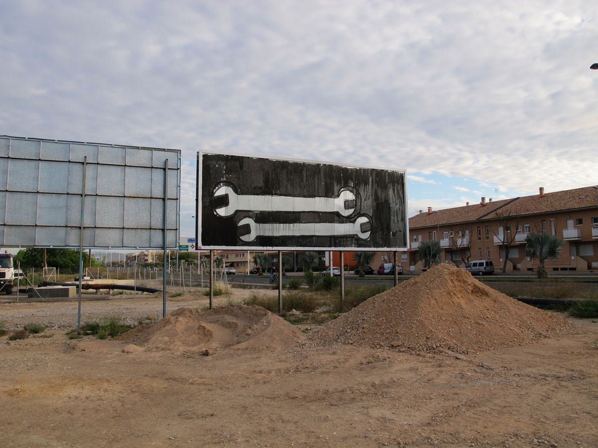 Black and white billboard painted as a large adjustable wrench lying horizontally, black paint dripping down the sign, dirt mounds in front.
