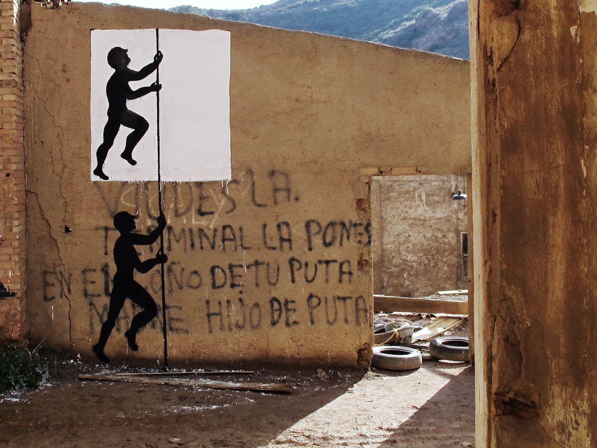 Two identical black silhouette figures climbing a vertical rope on a ruined building wall, one framed by a painted white rectangle, spray-painted graffiti text covering the rest of the wall.
