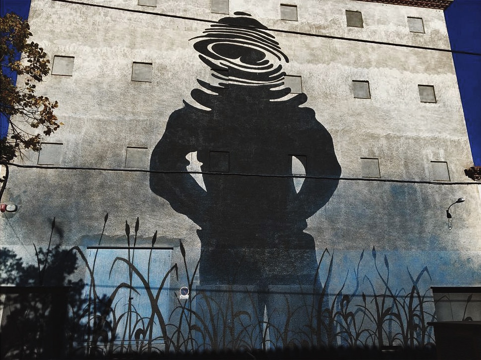 Large black silhouette mural on a building facade of a man standing with hands on hips, his head replaced by concentric rippled circles like water rings, painted reeds at the base of the wall.