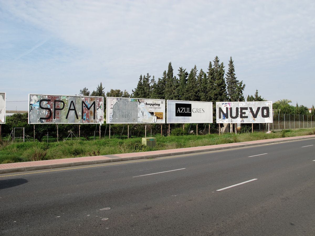 Row of four roadside billboards along a street, their surfaces stripped and painted with single black words reading 'SPAM,' a torn poster, 'AZUL GRES,' and 'NUEVO.'