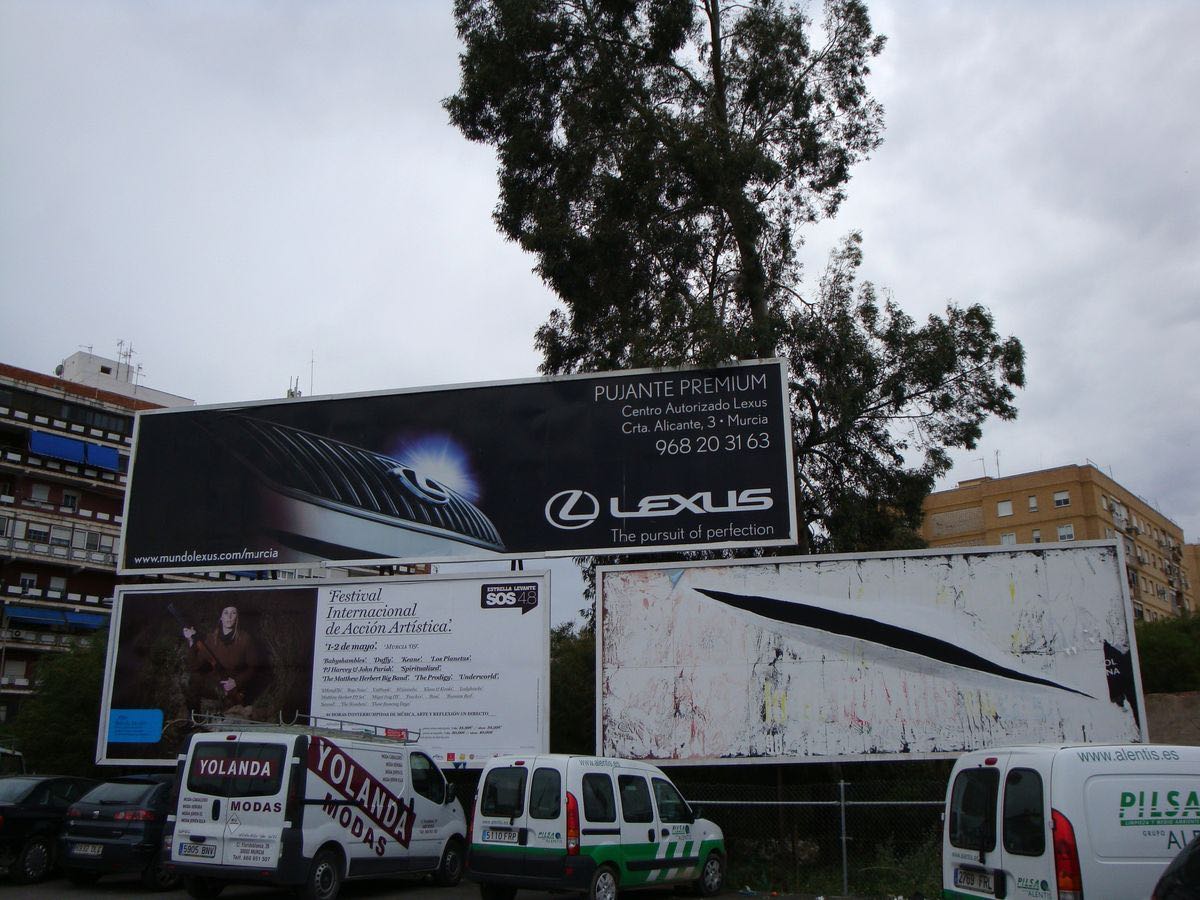 Row of advertising billboards beside a road, the lower right panel painted with a single diagonal black slash cutting across an otherwise blank white surface, echoing Lucio Fontana's slashed canvases, delivery vans parked below.