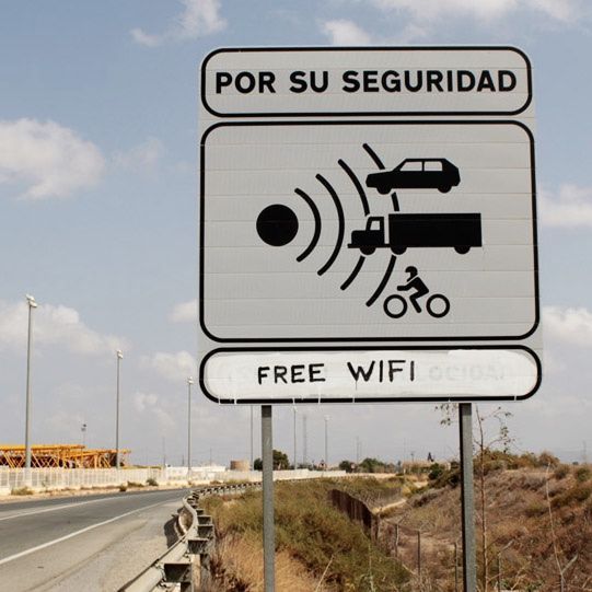Road speed-camera warning sign showing icons of a car, truck, and cyclist beside a radar symbol, with the words 'Free wifi' spray-painted beneath the official text.