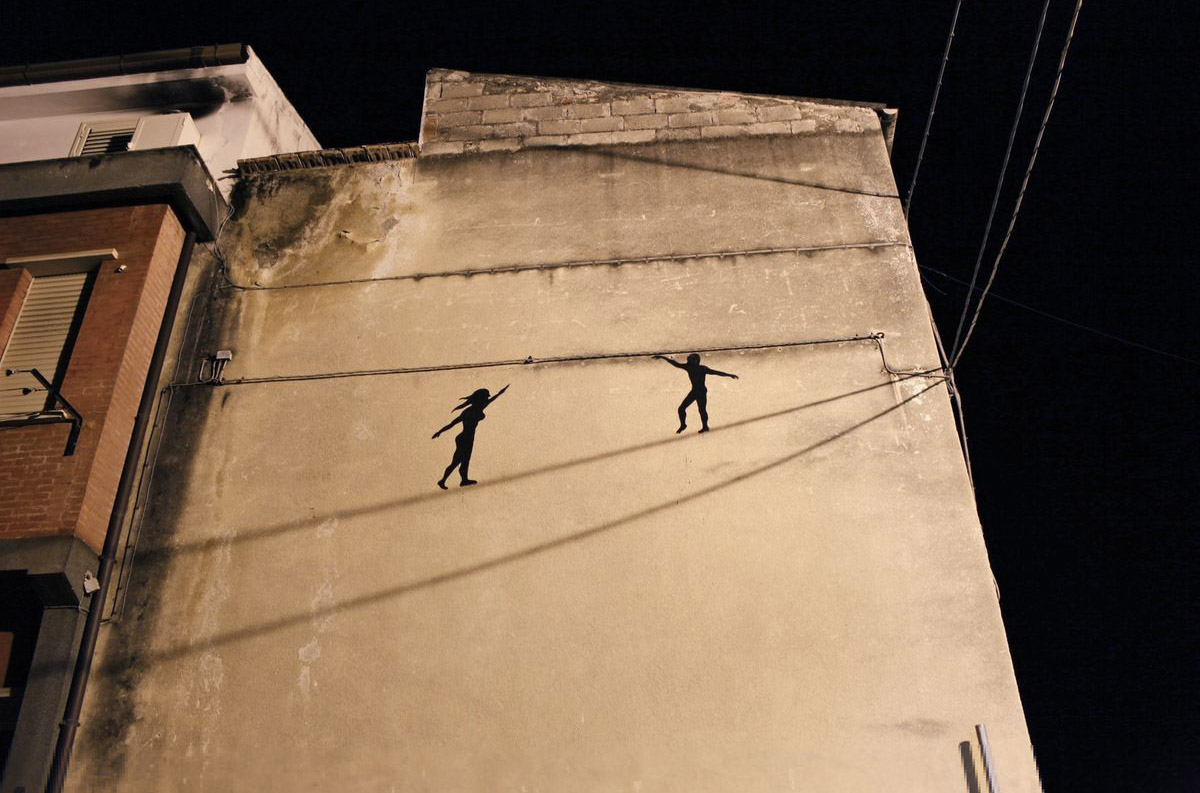 Two-photo day and night comparison of a building corner mural showing two black silhouette tightrope walkers balanced on overhead wires, parked cars below in both shots.