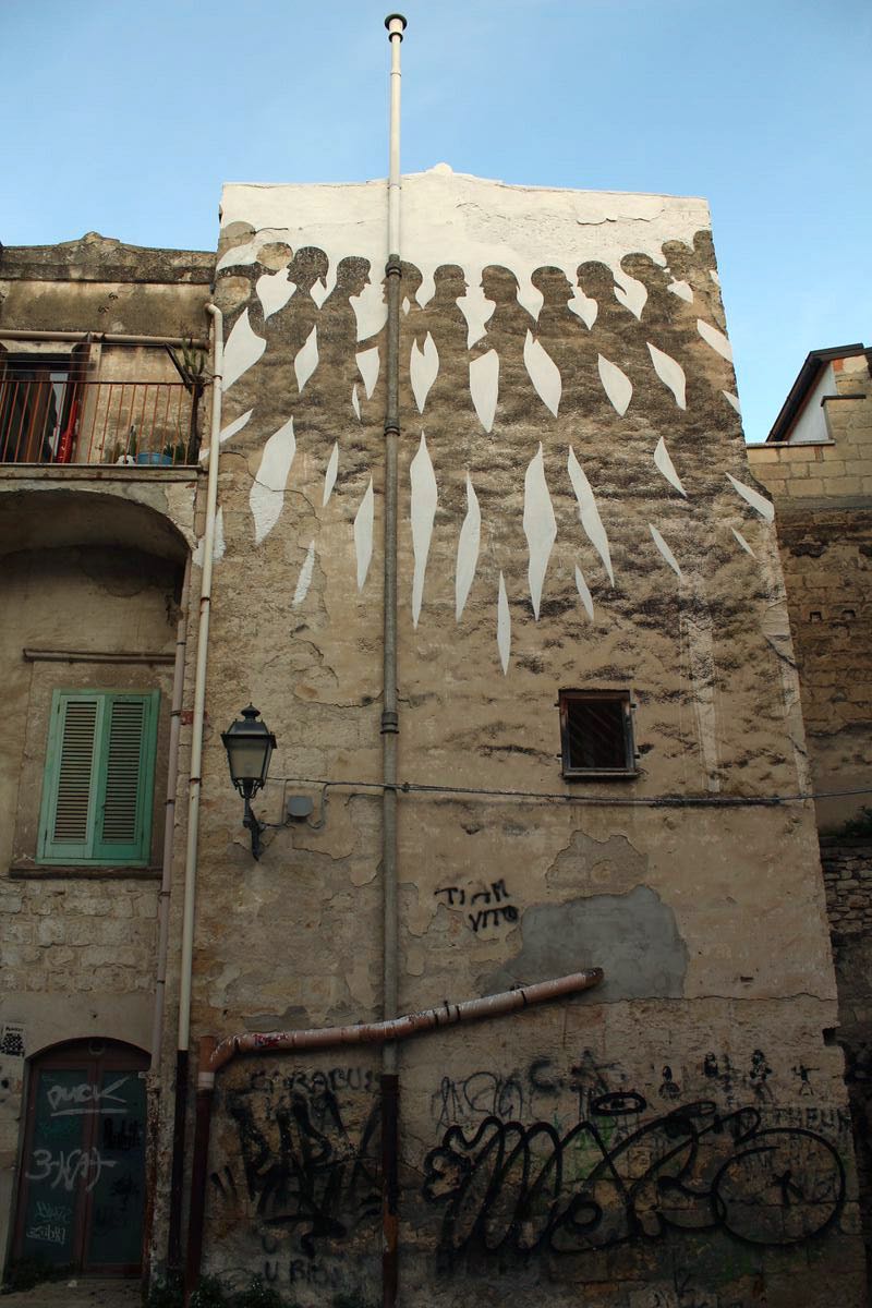 Black-and-white mural on a building facade showing a row of profile figures marching in a chain, each linked by a leaf-shaped white gap, above a window and a drainpipe.