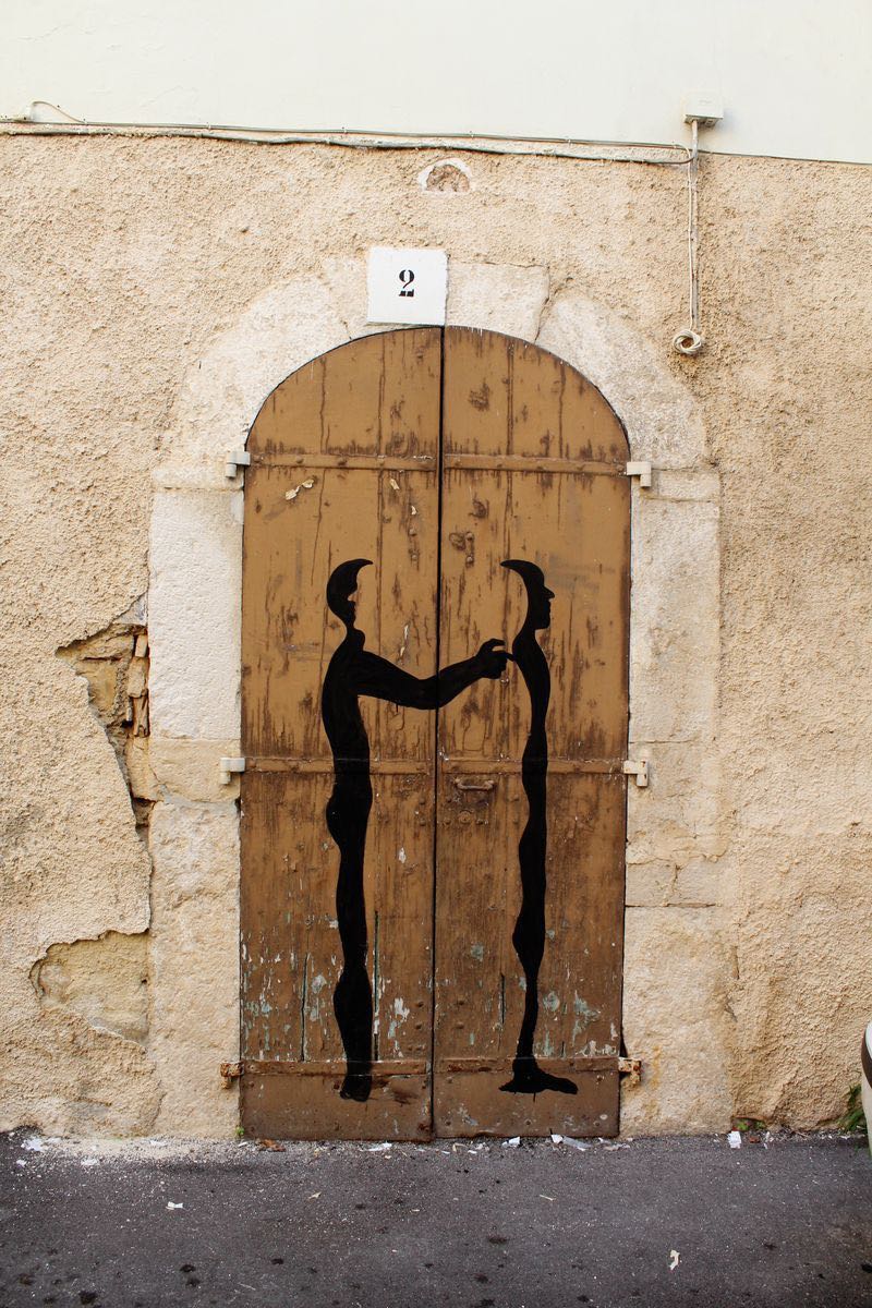 Two identical black silhouette figures in top hats reaching to shake hands, painted on a weathered wooden double door beneath a number 2 sign, set in a stone archway.