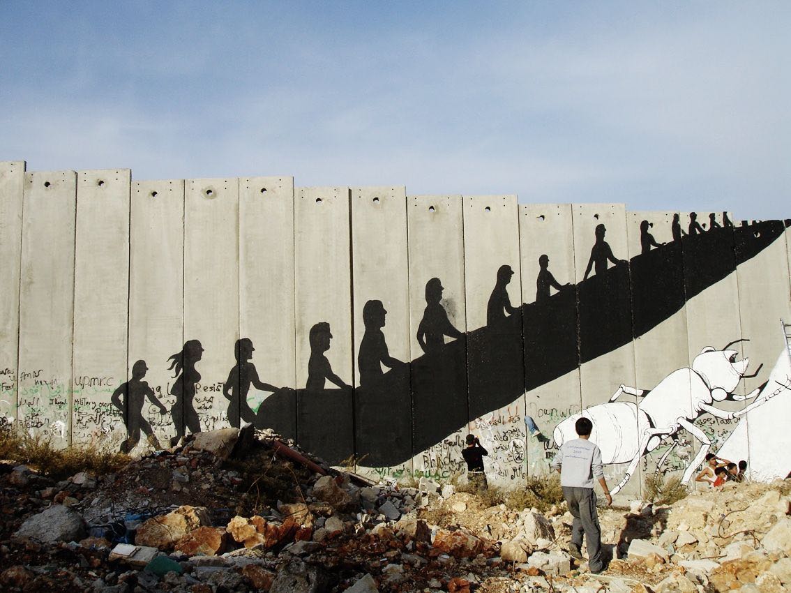 Row of black silhouette figures ascending a diagonal escalator-like line painted across a concrete separation wall, a white livestock animal painted at the base, rubble and children in the foreground.