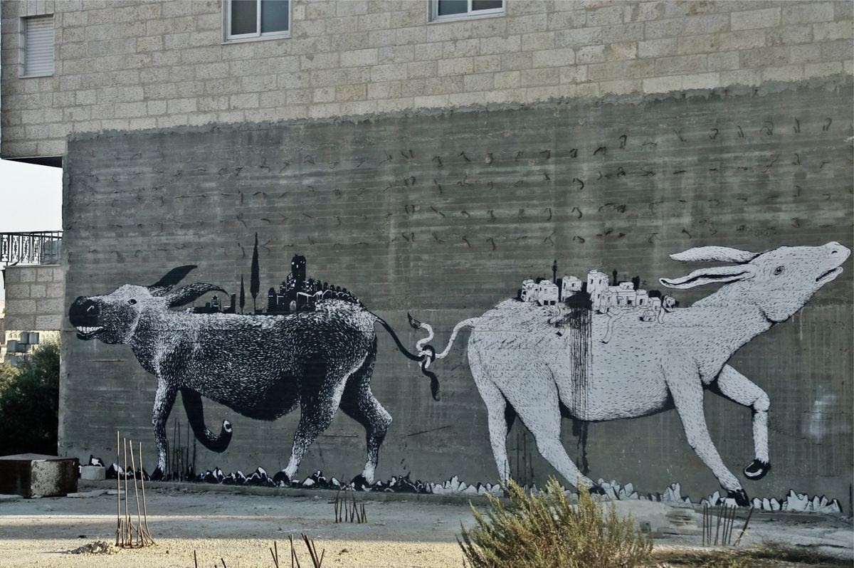 Two black-and-white donkeys facing away from each other on a concrete wall, each carrying a miniature hilltop town on its back, their tails intertwined with a snake between them, birds scattered across the wall.