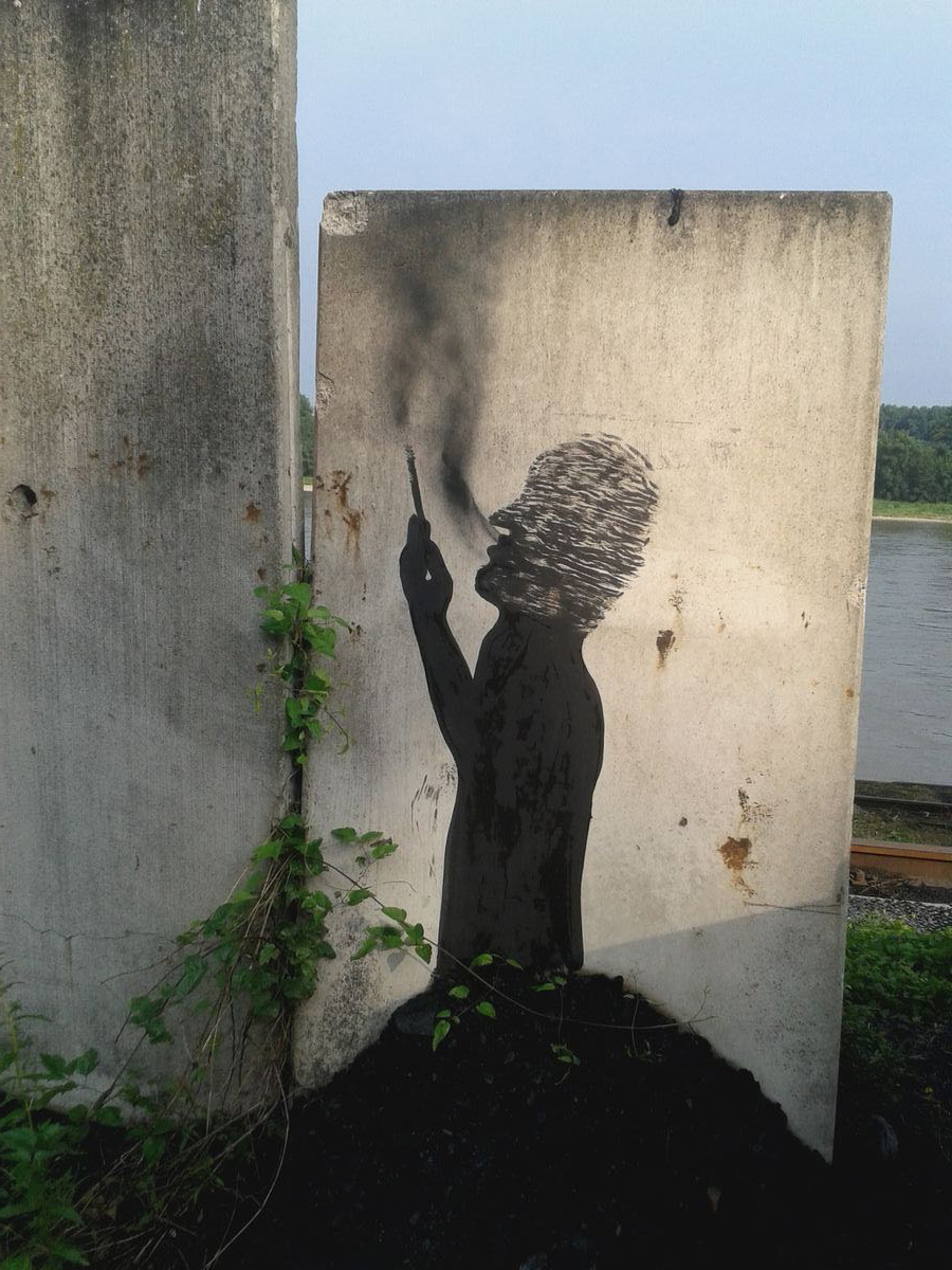 Black silhouette of a seated figure smoking a cigarette, exhaling a trail of smoke, painted on a concrete slab beside a low wall, framed by climbing green vines.