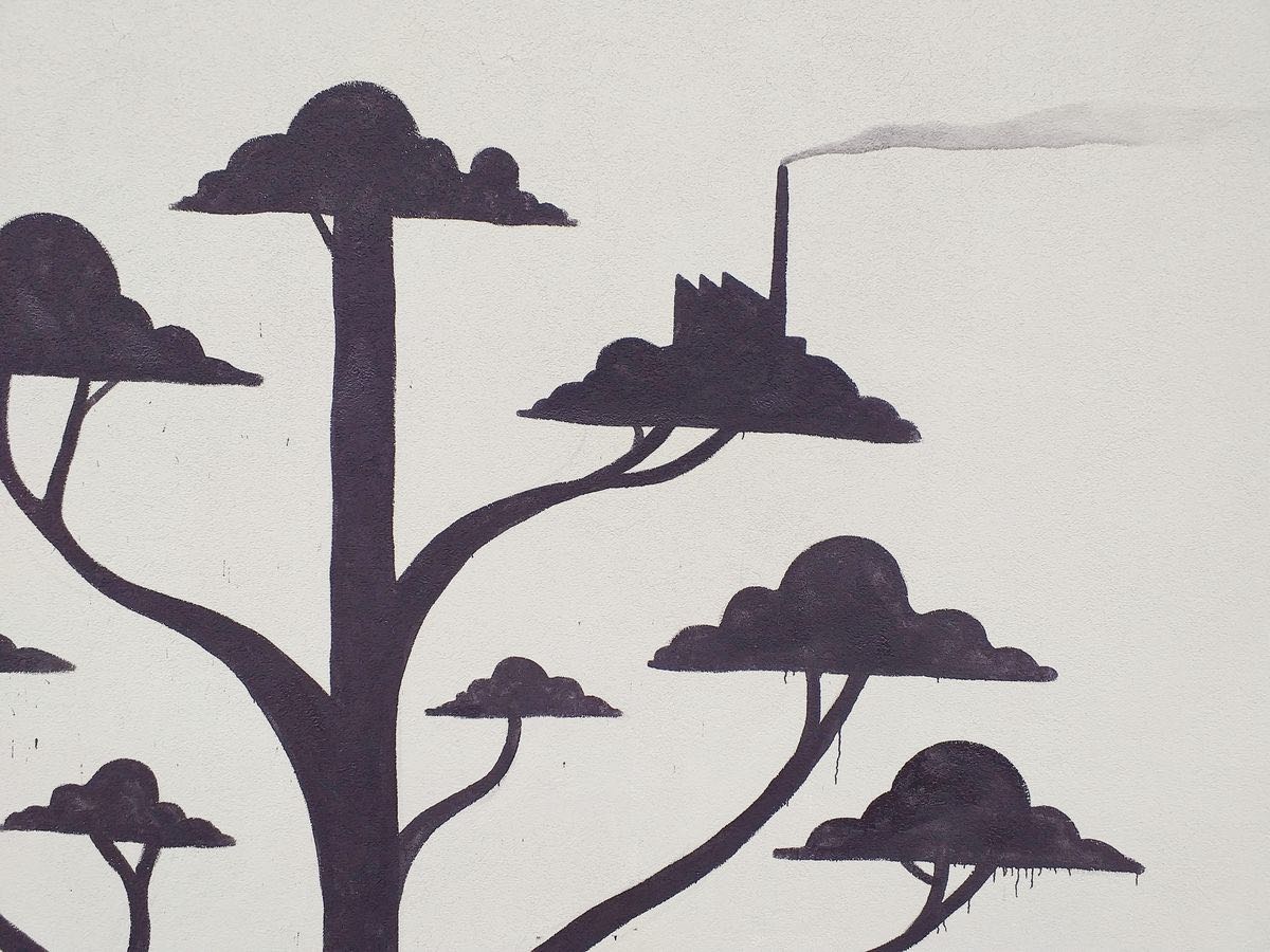 Detail of black silhouette trees with cloud-shaped canopies, one canopy shaped like a small factory with a chimney releasing a trail of smoke, painted on a plain wall.