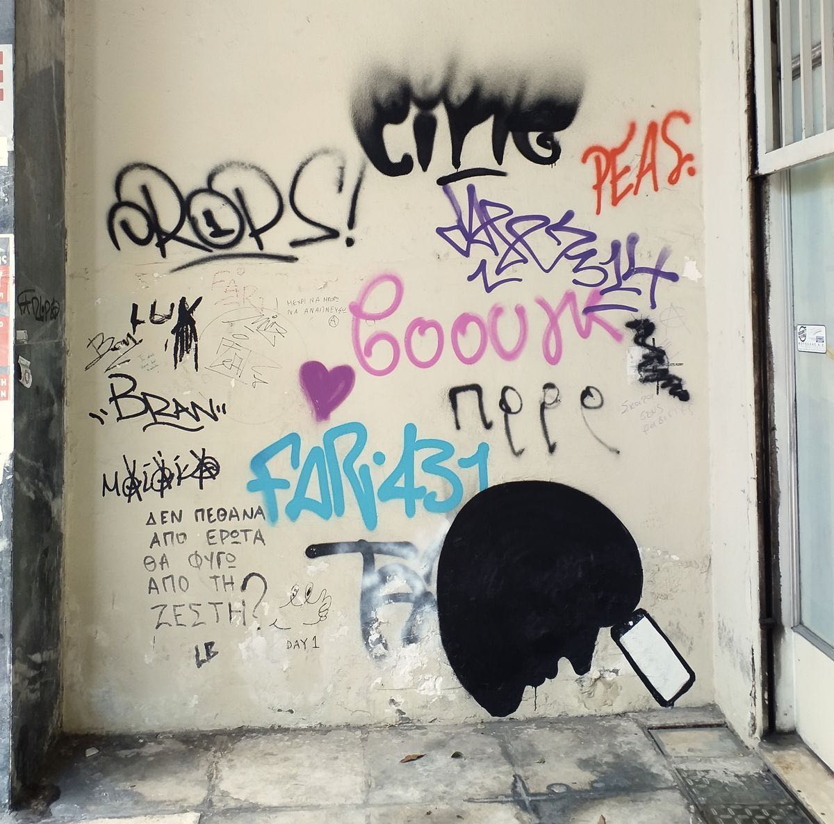 Black silhouette of a round head holding a smartphone up to its face, painted on a plastered wall covered in colorful spray-painted graffiti tags and handwritten notes.