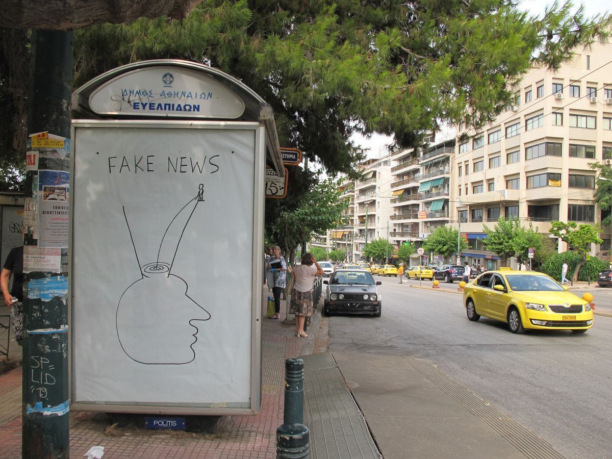 Outline drawing of a head with two V-shaped antennae rising from the top, a tiny figure balanced on one antenna tip, drawn in black marker on a bus shelter poster above the words Fake news.