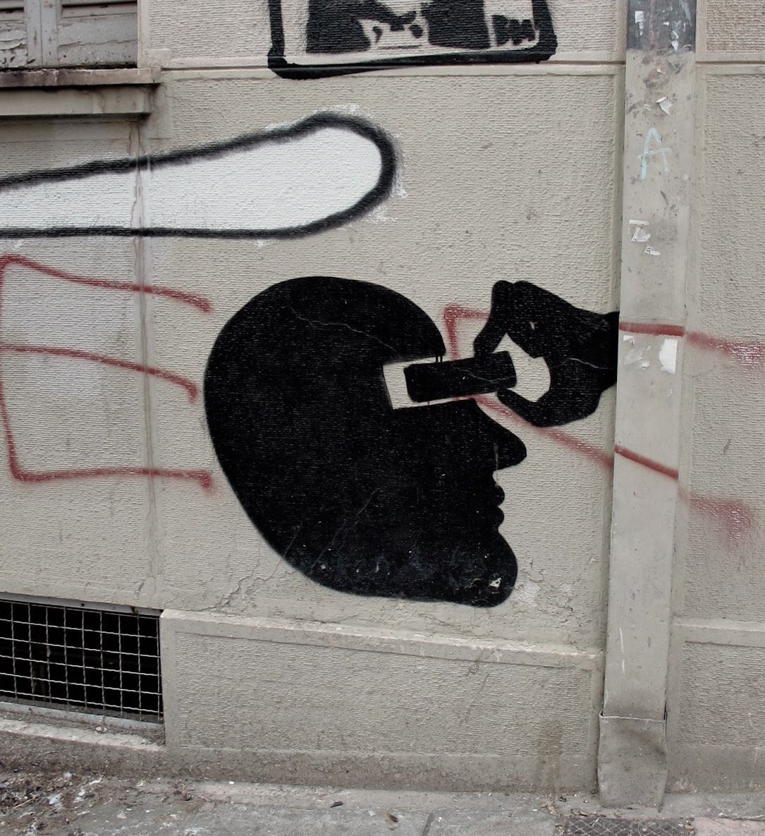 Black silhouette of a head in profile with a hand inserting a USB memory stick into a slot cut into the skull, painted on a textured wall beside red and white graffiti marks.