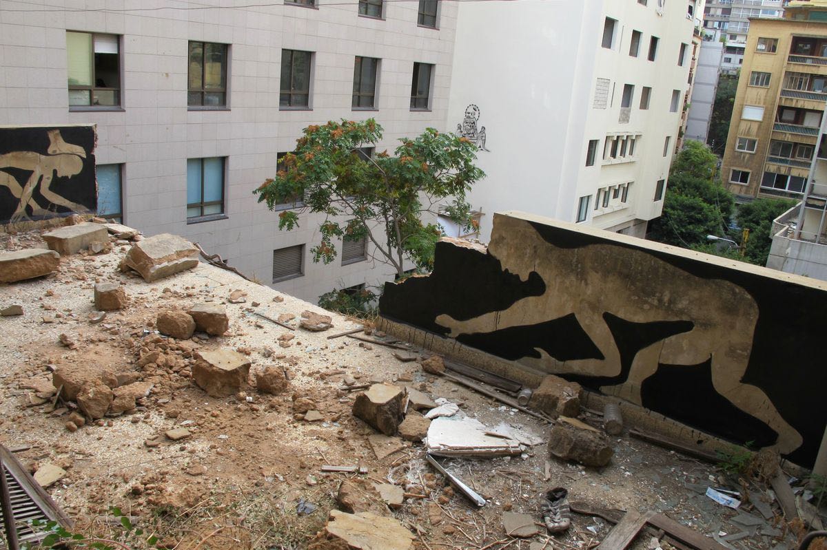 Aerial view from a rubble-covered rooftop showing two black silhouette murals of running or leaping figures painted on adjacent building walls, with a distant third silhouette visible on another building in the background.