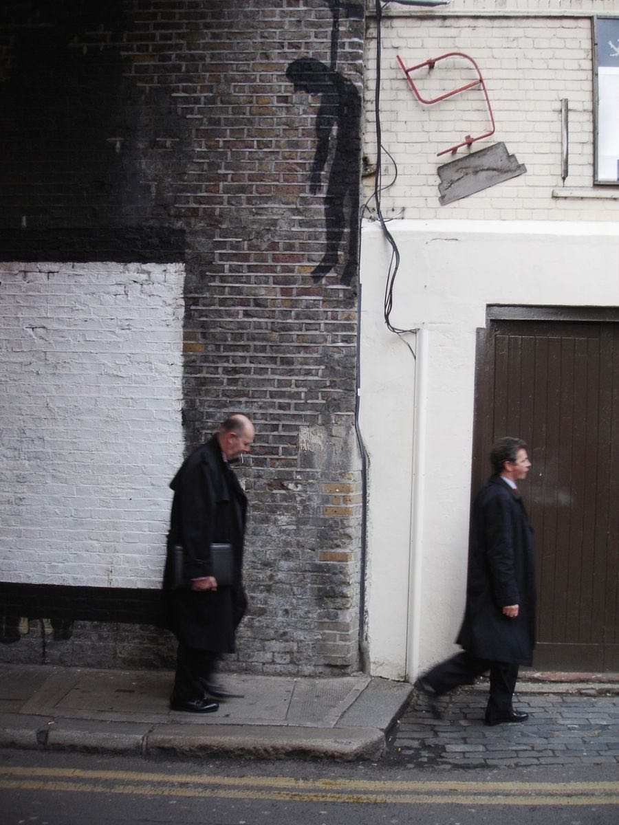 Black silhouette of a bent, downcast hanging figure painted high on a brick wall corner, two men in dark coats walking past below, red metal fixtures mounted on the wall.