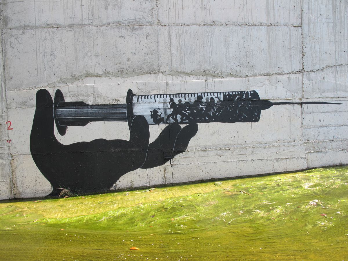Black silhouette mural of a hand holding a large syringe, with a crowd of running figures depicted inside the syringe barrel, painted on a concrete wall above a green algae-covered canal.
