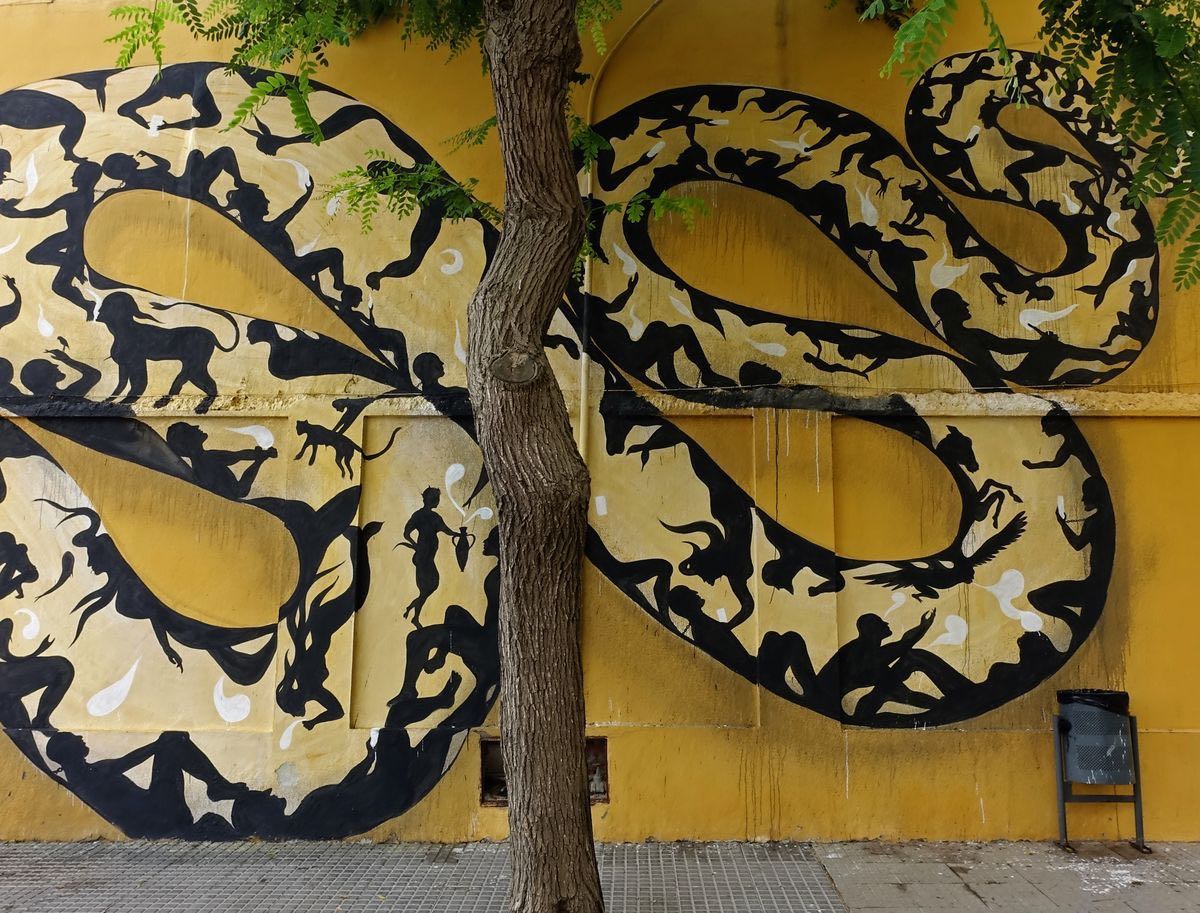 Wide view of two large coiled loops of the snake mural wrapping around a tree trunk, filled with black silhouettes of people and animals on the yellow building wall, a litter bin beside it.