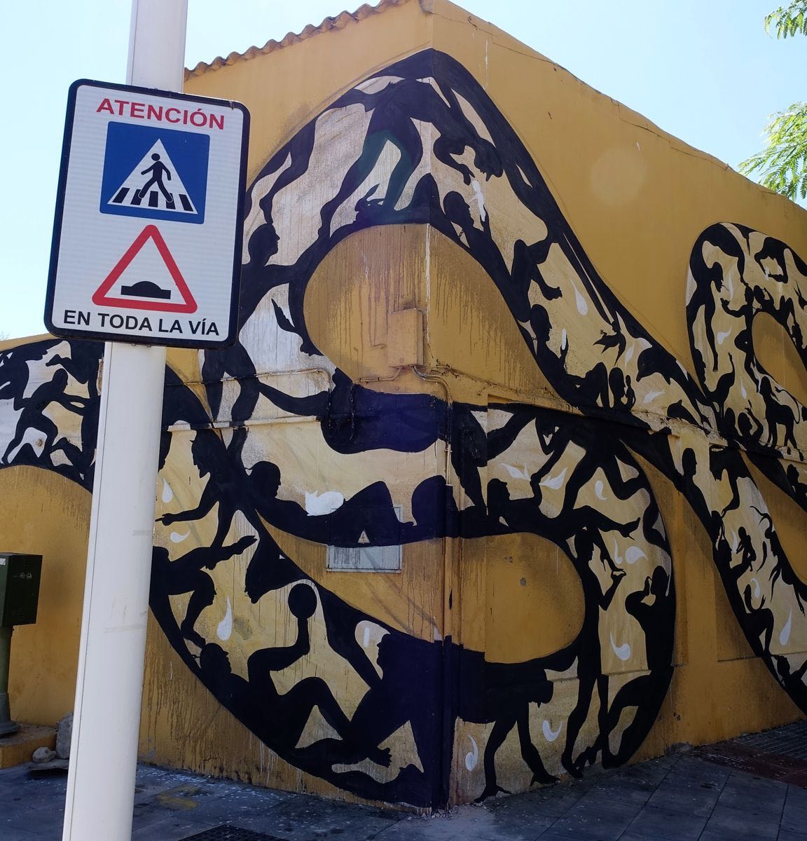 Corner view of the snake mural's coiled body filled with silhouetted figures climbing and playing, a pedestrian crossing and speed bump warning sign on a pole in front.