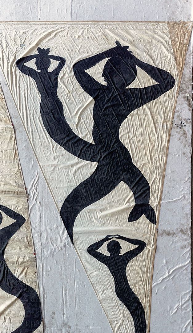 Close-up of a triangular sail with black silhouette figures of mermaids with raised arms and crowned heads painted on canvas, stains visible on the fabric.